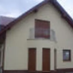 Otynkowany dom jednorodzinny z widoczną elewacją w jasnym odcieniu, brązowe okna i drzwi, balkon z metalową barierką, dach pokryty czerwoną dachówką, pochmurne niebo w tle.